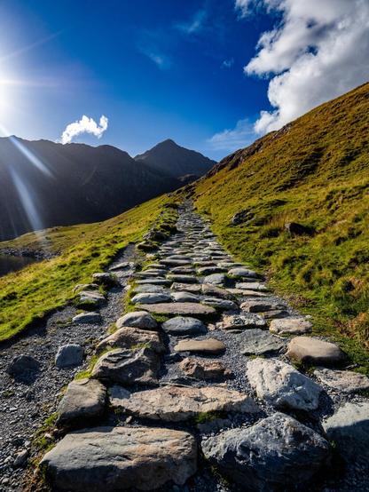 A wide stone-flagged path built from interlocking boulders leads upward on a steep mountain ridge cutting through a vibrant grassy slope. Up ahead dark mountain peaks rise in the distance under a blue sky with scattered clouds. Some sun rays shine on the left side of the image.