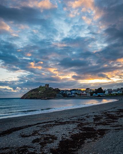 A wide-angle shot of a pebble beach in Criccieth, Wales at sunset. The ocean washes onto the shore and the beach leads to an outcropping in the distance with the silhouette of Criccieth Castle standing at the top of a grassy hill. The houses of the coastal town can be seen on the shore under a cloudy, dramatic blue and orange sky.