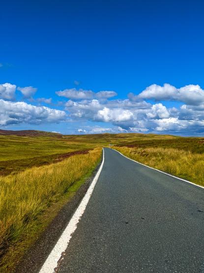 A scenic winding road cuts through the open green and golden hills of a moorland landscape under a bright, cloud-dotted blue sky.