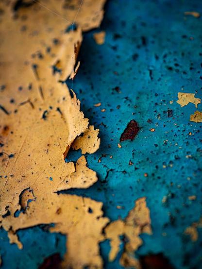 A macro photo of a heavily textured layer of yellow paint cracking and curling up off of a bright turquoise layer on metal. The blue coloured metal has pitting and rust specks.