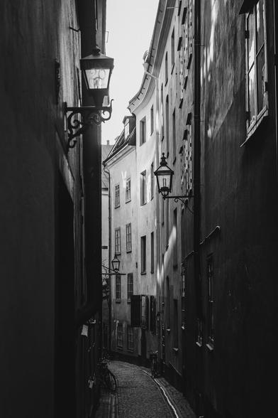 A vertical monochrome photograph of a narrow alley paved with cobble stones, On both sides of the alley are a few stories tall old buildings with plastered walls. In the foreground the alley is quite dark while further back daylight comes down and illuminates the buildings to the right where the alley makes a bend and disappears. Reflected sunlight casts a few brighter spots on the walls. Old style lamps, some which have house numbers on them are set in the walls.

A few bicycles line the alley, which otherwise is empty.