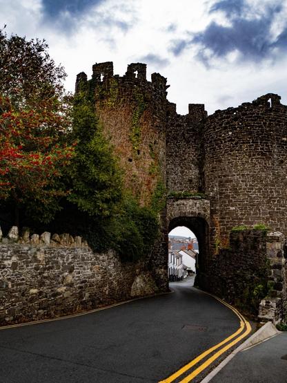 A winding two-way asphalt road with double yellow lines leads through a medieval stone archway. A large, dark stone tower covered in ivy stands on the left, next to green trees with red berries. A downhill street with white terraced houses can be seen through the archway under a cloudy, overcast sky.