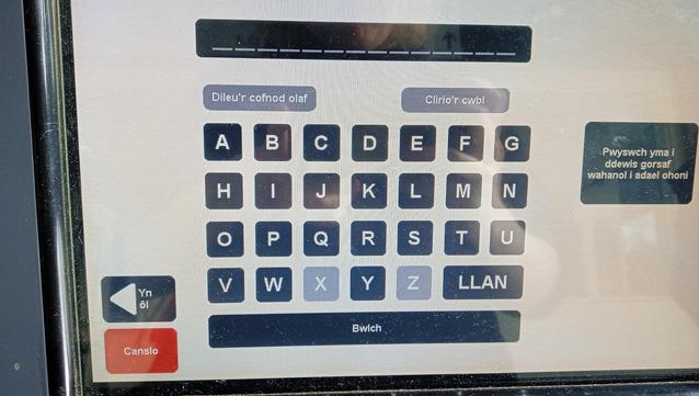 a photo of a transport for wales ticket machine, it is on the destination selector screen, there is an alphabetically arranged 7x4 keyboard button grid, on the bottom right of the grid is a two wide button which says "LLAN", other buttons for going back to the previous screen and cancelling are on the left
