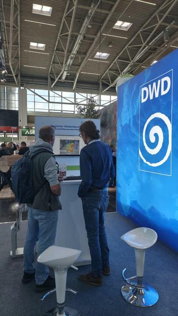 Ein Foto zeigt, wie ein DWD Mitarbeiter einem Messebesucher der Interforst den Bodenfeuchteviewer des DWD auf einem Bildschirm zeigt. Quelle: DWD