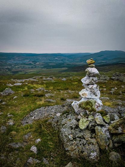 A tall, narrow rock cairn built from stacked lichen covered stones stands balanced on a larger rock formation. The ground is covered in green and yellow grasses. In the background, a wide valley sits beneath a colourless, overcast sky.