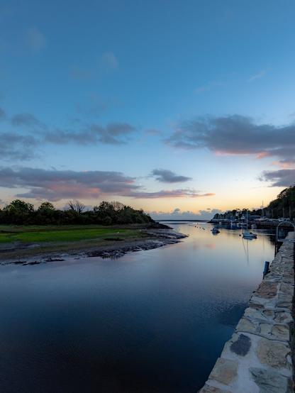 Scenic view from a stone harbor wall looking out over Porthmadog Harbour at dusk. The calm water reflects a soft blue sky with pink clouds and golden light on the horizon. A small grassy island with trees sits to the left of the image, while small sailing boats are docked along the right side near the town.